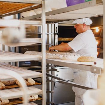 Im Vordergund Regale mit frisch gebackenem Brot. Im Hintergrund steht der Bäcker und bereitet Brote fürs Backen vor.