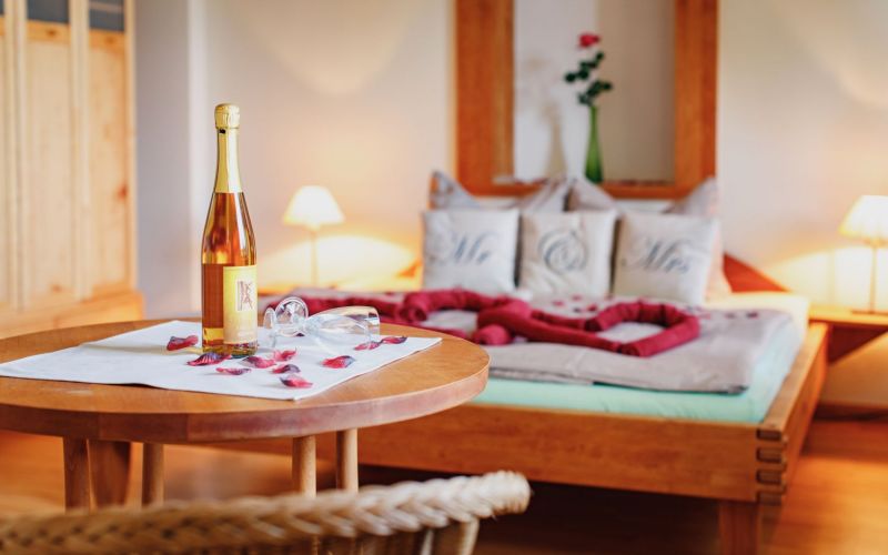 Im Vordergrund ein Tisch dekoriert mit Rosenblüten und einer Flasche Sekt. Im Hintergrund ein Bett mit Herzkissen.