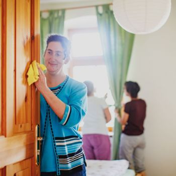 Eine lächelnde Frau wischt eine Holztür ab. Im Hintergrund zwei Frauen bei der Raumpflege.