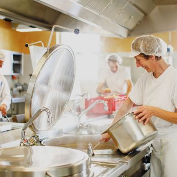 Zwei Männer und eine Frau in weißer Arbeitskleidung bei der Arbeit in der Großküche. Die Frau im Vordergrund schöpft Suppe aus einem großen Topf.